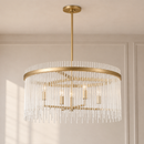 A modern drum chandelier with a brushed gold finish and clear glass rod drops, suspended from a matching gold ceiling canopy in a neutral room setting.