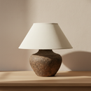 A table lamp with a textured, rustic brown ceramic or stone-look base and a tapered off-white fabric drum shade, shown against a plain white background.
