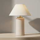 A table lamp with a textured cream cylindrical base and a matching cream tapered fabric shade, illuminated with a warm glow. A small gold metal accent is visible at the top of the base.