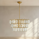 A gold and clear glass tiered chandelier hanging from a gold ceiling canopy against a beige wall with soft window light casting shadows.