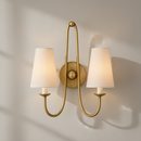 A double-arm wall sconce with a warm gold/brass-finished metal frame and two tapered off-white fabric shades, mounted on a beige wall with a small plant visible in the lower right corner.