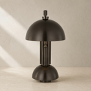 A modern table lamp with a matte black dome shade and matching cylindrical base, shown on a light textured surface against a neutral beige wall with soft diagonal shadows.