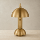 A modern table lamp in a brushed brass or gold finish, featuring a dome-shaped shade, ribbed central column, and matching dome base, set against a neutral textured beige background.