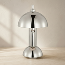 A polished chrome table lamp with a dome-shaped shade and matching base, standing on a light beige surface against a textured beige wall.