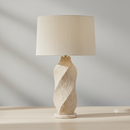 A table lamp with a textured, twisted beige stone base and a matching beige linen drum shade, set against a neutral gray background.
