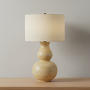 A table lamp with a textured, double-bulb gourd-shaped base in a light natural wood finish, topped with a cylindrical drum shade in a warm cream fabric.