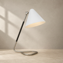 A modern table lamp with a white conical shade, a curved brushed silver metal base, and a black textured stem, shown on a light stone surface against a textured beige wall with soft window shadow patterns.