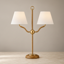 A two-light table lamp with a polished gold-finished metal frame, featuring textured rope-style arms, a circular top loop, and a round woven base. It has two tapered off-white fabric shades, shown lit on a light stone surface against a textured beige wall.